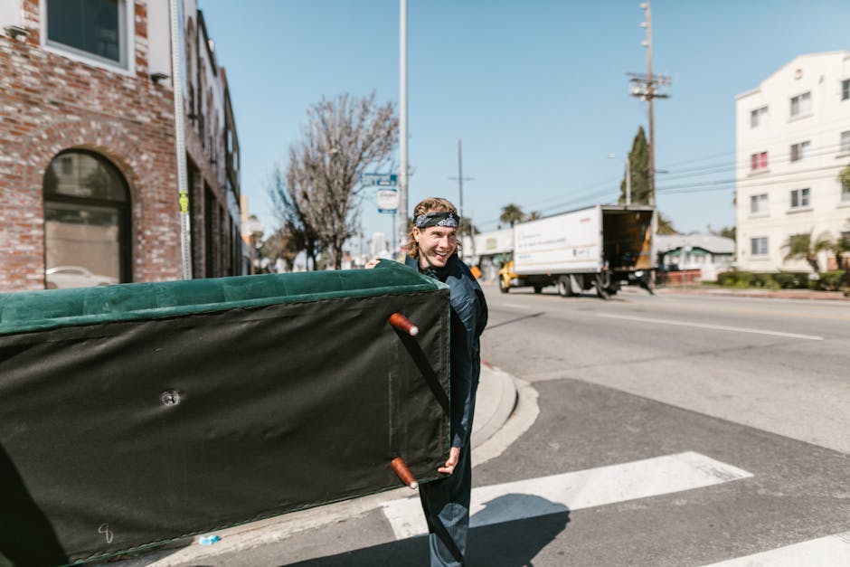 A person wearing a protective headband is standing on a sidewalk and carrying a large, rectangular moving box covered with green and black fabric, secured with straps for stability during the home relocation process. The individual is smiling and dressed in casual clothing, positioning the box upright as part of a furniture transport or packing and moving activity. In the background, there is a busy street scene with a white moving truck parked with its rear open, a cargo vehicle on the road, and several multi-storey buildings, including a brick structure to the left and a white apartment building to the right. The scene is set in daylight under clear skies, with some trees and utility poles visible, indicating an urban environment suitable for house removals. This image illustrates the loading process involved in house removals, highlighting packing materials, equipment such as straps, and the movement of furniture or large items for a shift to or from premises, consistent with services offered by Removals Edgware for shopping area moves in Edgware.
