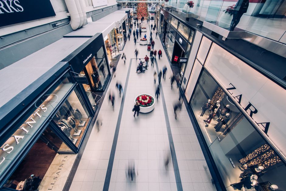 A high-angle view of an indoor shopping mall corridor with multiple retail stores on either side, including visible storefronts for Saks Fifth Avenue and Zara, with illuminated display windows showcasing mannequins dressed in clothing. The polished tiled floor reflects the overhead lighting, and a central circular flower planter with red and white flowers is positioned along the walkway. Numerous blurred shoppers walk through the space, some carrying shopping bags, indicating movement and busy retail activity typical of a shopping area. The scene suggests a vibrant environment where furniture, packaging materials, or other household items may be transported or relocated during a house move, and professionals like Removals Edgware are experienced in managing such logistics during home relocation or packing and moving processes. The image emphasizes the importance of careful space management and the smooth loading of items, akin to the logistics involved in moves and transportation within shopping centers.