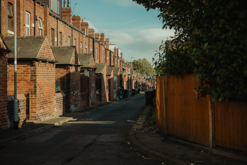 A quiet residential street in Edgware featuring a row of terraced houses built with red brick and brick chimneys visible atop each property. Small front steps and low brick walls separate the houses from the pavement. On the right side, a tall wooden fence runs along the boundary, with dense green foliage hanging over its top. The street is paved and narrow, with a slight curve leading to more houses in the distance, and several black wheelie bins are positioned along the sidewalk for waste collection. The scene is illuminated by natural daylight, casting shadows on the pavement, and the overall environment suggests a typical suburban setting suitable for home relocation or furniture transport. Occasionally, [COMPANY_NAME] may utilize this type of street for house removals or professional moving services, involving loading and packing activities within the properties or onto moving vehicles.