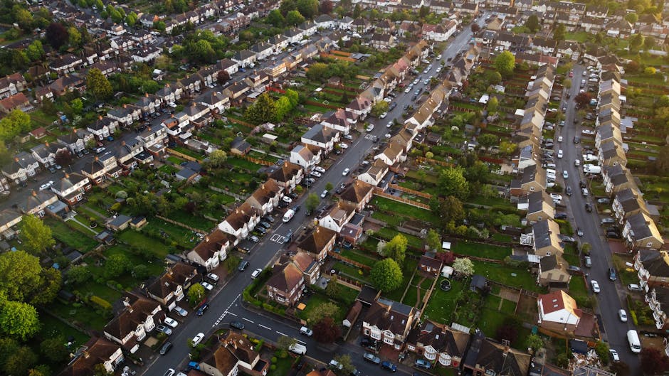 Aerial view of a residential neighbourhood showing rows of terraced houses and semi-detached homes in Edgware, with streets lined with parked cars. The gardens at the back of the houses contain trees and green lawns, some with visible fencing and outdoor furnishings. The scene captures the organized layout of the area, with the roads connecting the properties and a mix of vehicles including cars and vans parked along the streets, highlighting a typical setting for a house relocation or home moving process. The lighting indicates a daytime setting with natural daylight illuminating the scene, making it suitable for illustrating furniture transport, packing and moving activities, or the loading process conducted by Removals Edgware, specialists in house removals in the Hale Lane HA8 area.