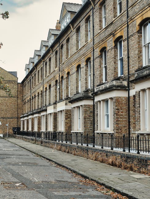 A quiet residential street in Edgware featuring a row of terraced houses built with red brick and brick chimneys visible atop each property. Small front steps and low brick walls separate the houses from the pavement. On the right side, a tall wooden fence runs along the boundary, with dense green foliage hanging over its top. The street is paved and narrow, with a slight curve leading to more houses in the distance, and several black wheelie bins are positioned along the sidewalk for waste collection. The scene is illuminated by natural daylight, casting shadows on the pavement, and the overall environment suggests a typical suburban setting suitable for home relocation or furniture transport. Occasionally, [COMPANY_NAME] may utilize this type of street for house removals or professional moving services, involving loading and packing activities within the properties or onto moving vehicles.