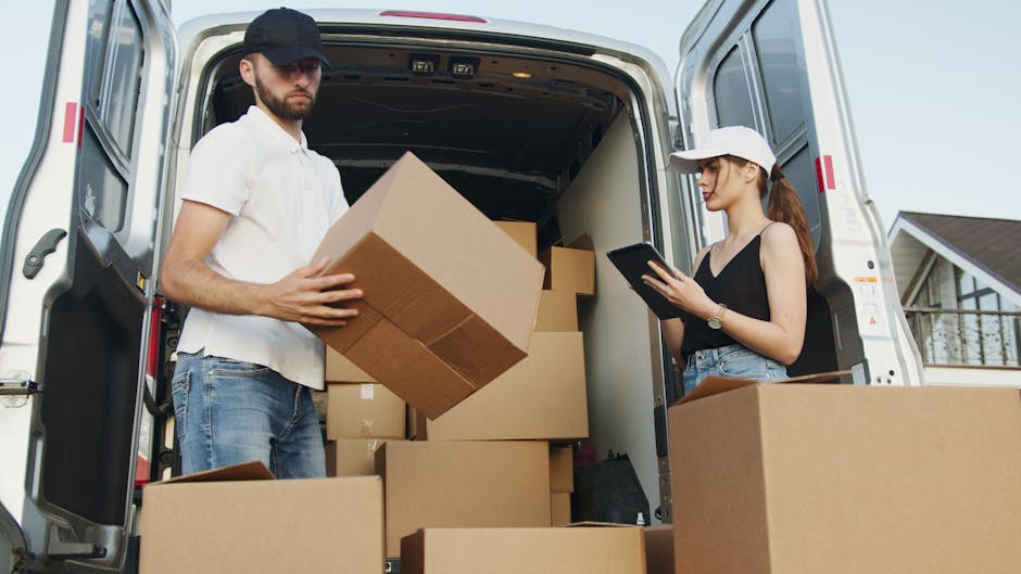 Two individuals are loading and organizing cardboard boxes inside the open back of a white moving van, parked outside a residential property on a paved driveway with a house visible in the background.