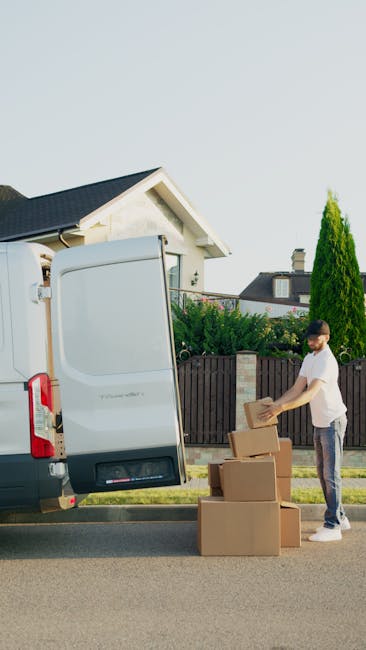 Two individuals are loading and organizing cardboard boxes inside the open back of a white moving van, parked outside a residential property on a paved driveway with a house visible in the background.